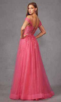 A woman models the Juliet JT2466A Sheer-Waist Long Prom Ball Gown, a pink off-the-shoulder dress with a lace-up open back and floral embellishments, standing before a gray background.