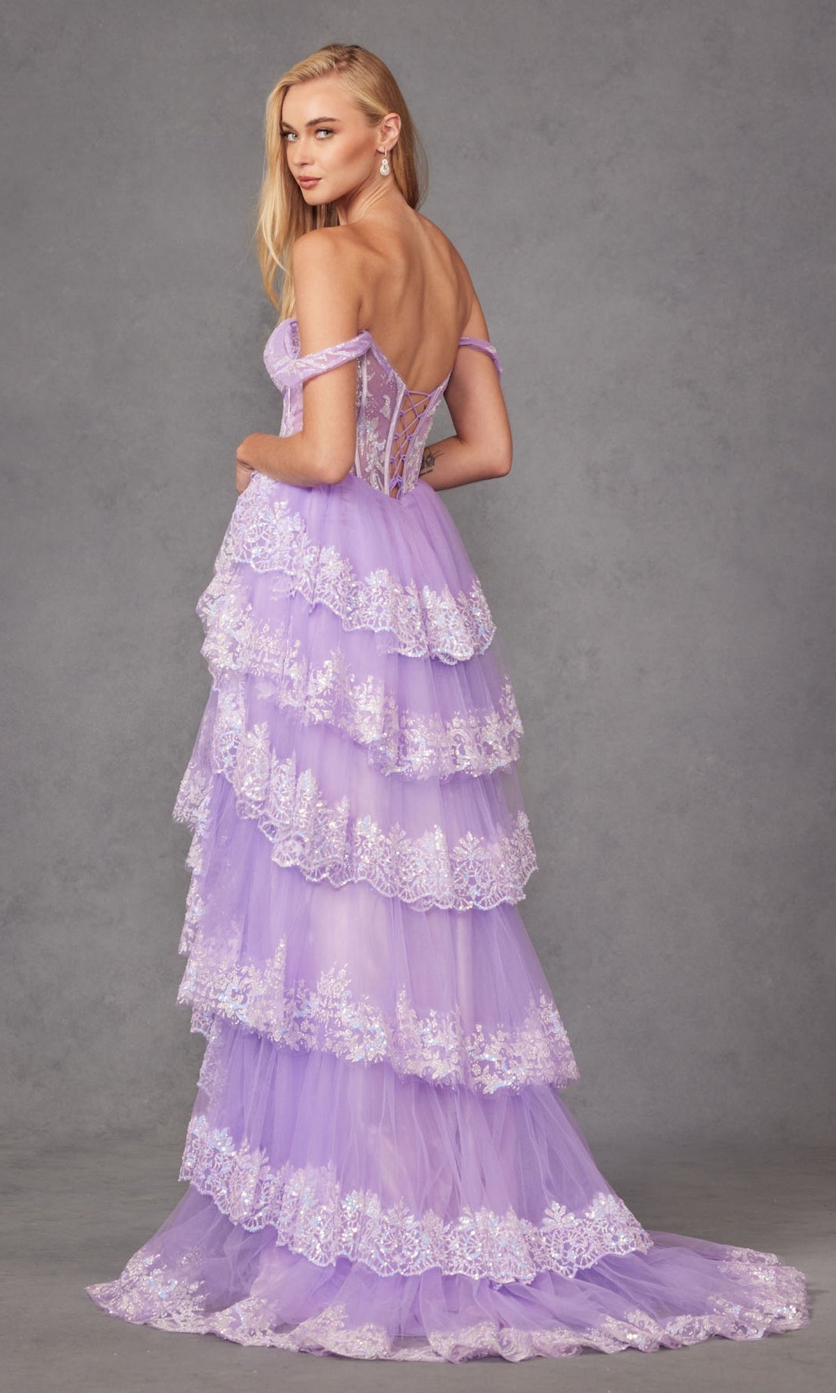 A woman models the Juliet JT2467K Off-the-Shoulder Ruffled Prom Dress in lavender, featuring tiered ruffles and white floral lace, against a gray background.