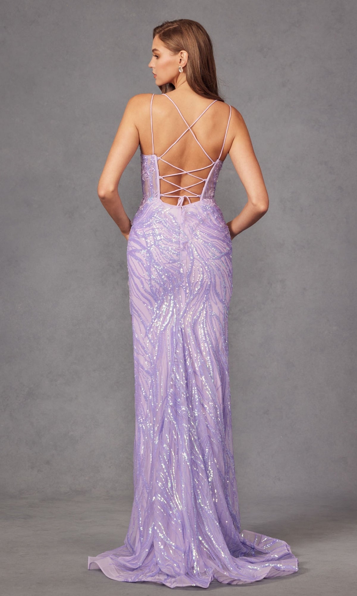 A woman models the Juliet JT2473R— a long lilac sequin prom dress with spaghetti straps and a lace-up back— against a plain gray background.