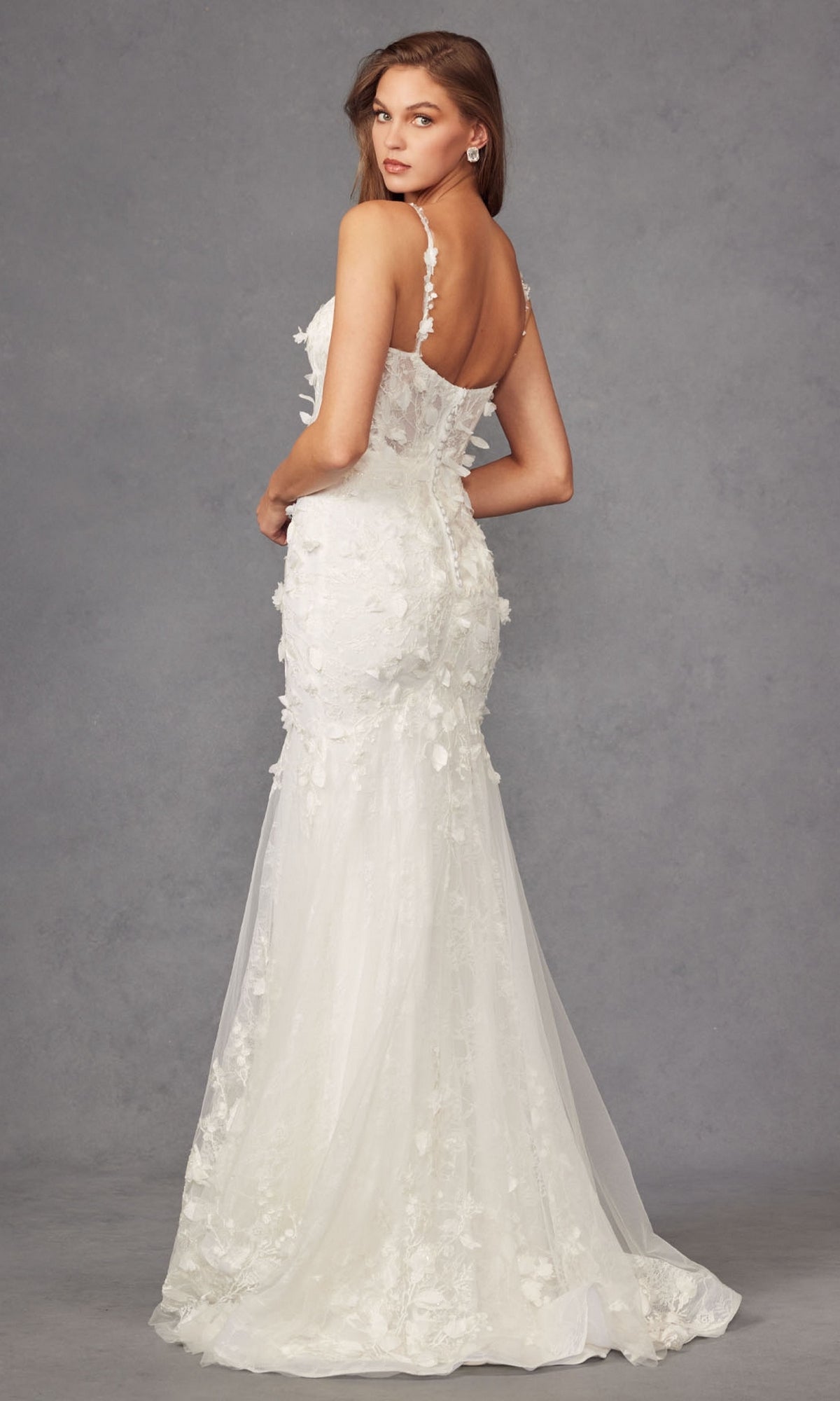 A woman models the Long Prom Dress JT2475UW by Juliet, a fitted, strapless off-white mermaid gown with floral appliqués, posing with her back slightly turned against a plain gray background.