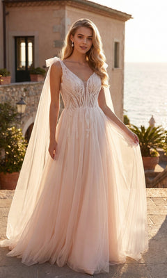 A woman stands outdoors near a stone building by the sea at sunset, wearing the Juliet JT2602CW long formal dress in nude, featuring sheer cape sleeves and an elegant A-line tulle skirt.