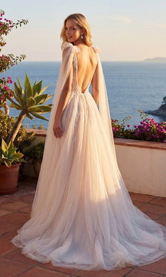 Wearing the Juliet JT2602CW, a long formal dress featuring a flowing, backless design and an elegant A-line tulle skirt, a woman stands on a terrace adorned with potted plants, gazing over the ocean at sunset.