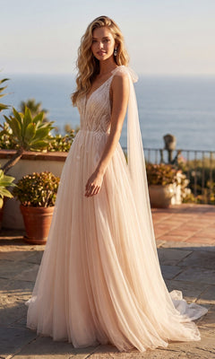 A woman stands outdoors in the Juliet JT2602CW long formal dress—a sleeveless, floor-length blush bridal gown featuring a sheer overlay and lace bodice. Surrounded by potted plants, she poses gracefully in the gown’s A-line tulle skirt, with the ocean shimmering in the background.