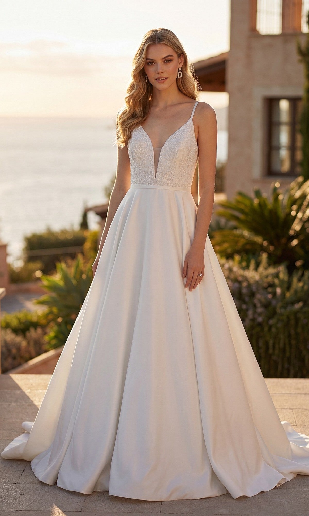 A woman wearing the Juliet JT2603CW long formal dress in off white—a modern bridal gown with a sleeveless design and deep V-neckline—stands outdoors near a building and greenery, with the ocean visible in the background.