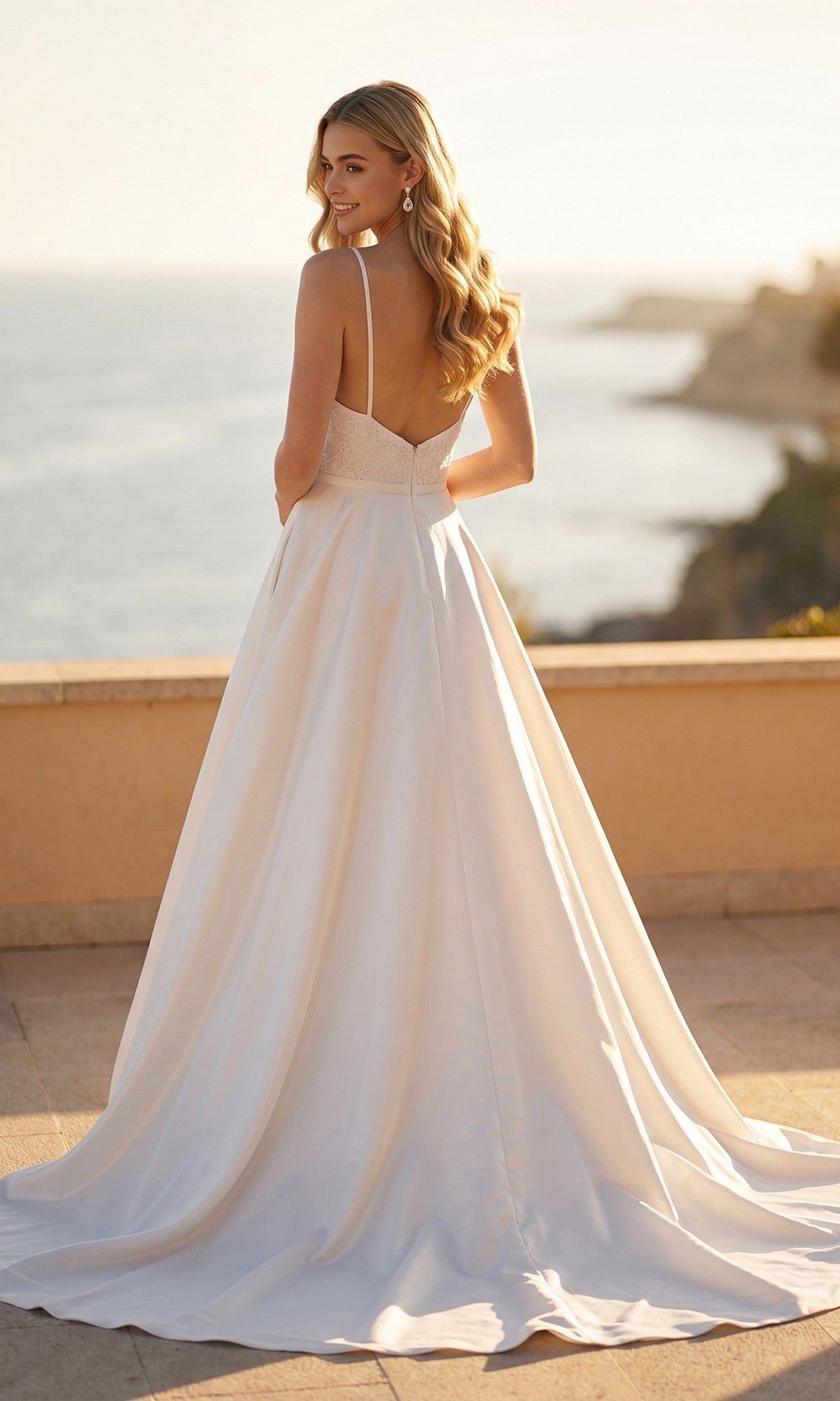 A woman wearing the Long Formal Dress: Juliet JT2603CW stands outdoors, facing away with her head turned, overlooking a sunlit coastal landscape.