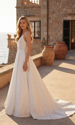 Wearing the Juliet JT2603CW long formal dress, a woman stands on a stone terrace adorned with large clay pots and overlooking the sea. Her sleeveless white satin A-line gown drapes elegantly, perfectly complementing the serene coastal setting.