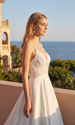 A woman wearing the Juliet JT2603CW long formal dress, featuring a white sleeveless lace bodice, stands on a terrace with trees, a building, and the sea in the background under clear skies.
