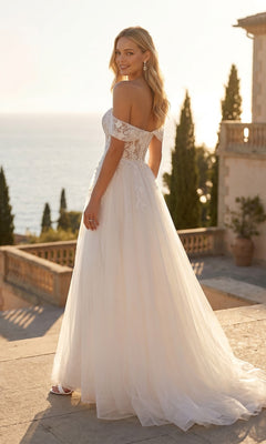 A woman in a Long Off-White Bridal Dress: Juliet 82606L with an off-the-shoulder illusion lace bodice and full tulle skirt stands on a stone terrace overlooking the sea.