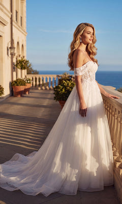 Long Off-White Bridal Dress: Juliet 82606L featuring an off-shoulder neckline, illusion lace bodice, and full tulle skirt, standing on a stone terrace overlooking the ocean.