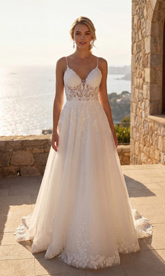 A woman stands outdoors on a stone terrace overlooking the sea, wearing the Juliet JT2633LW long formal dress in off white, a modern bridal gown with thin straps and elegant lace detailing.