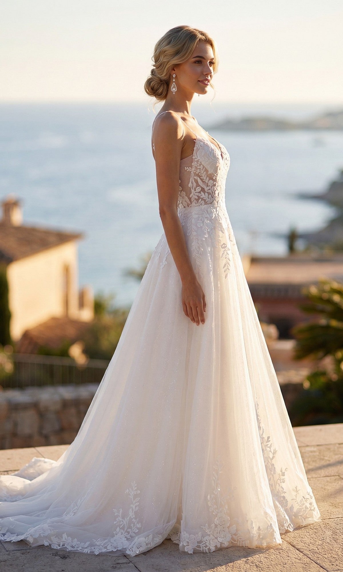 A woman stands outdoors near the coast at sunset, wearing the Juliet JT2633LW long formal dress—an off-white lace bridal gown—with buildings and the sea in the background.