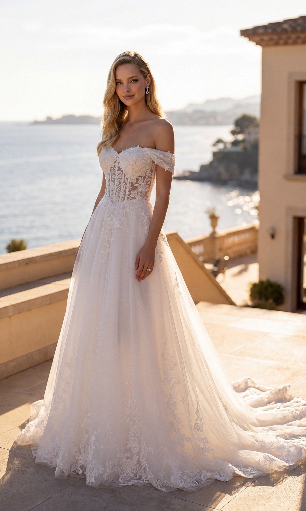 A woman in the Long Formal Dress: Juliet JT2634LW, an off-white, off-shoulder gown with lace appliqué, stands outdoors on a sunlit terrace overlooking the sea, with elegant buildings and a scenic coastline in the background—a stunning choice for a wedding reception dress.