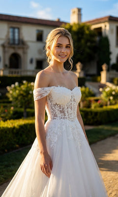 A woman wearing the Long Formal Dress: Juliet JT2634LW, an off-the-shoulder lace appliqué gown, stands outside in a garden with a large building in the background.
