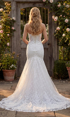 A woman wearing the Juliet JT2666SW long formal dress stands outdoors on stone pavement, facing a wooden gate surrounded by rose bushes. The strapless gown features delicate lace detailing.