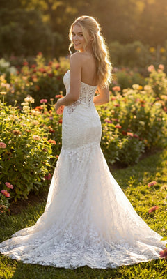 Wearing the Long Formal Dress: Juliet JT2666SW, a woman stands outdoors in a garden filled with blooming flowers, looking over her shoulder and smiling.