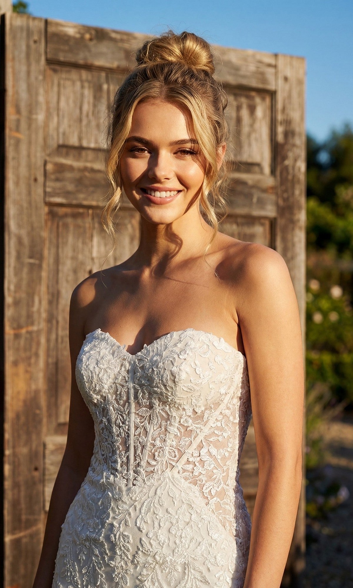Wearing the Juliet JT2666SW long formal dress, a woman stands outdoors in front of a wooden door, smiling at the camera as sunlight illuminates her face and hair.