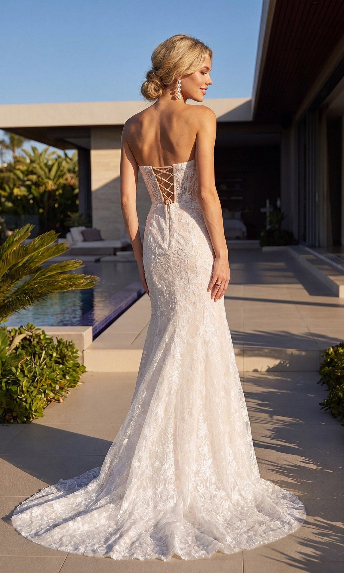 A woman in the Long Formal Dress: Juliet JT2667SW, a strapless lace floor-length gown, stands outdoors by a pool with her back to the camera, her hair styled in an elegant updo.