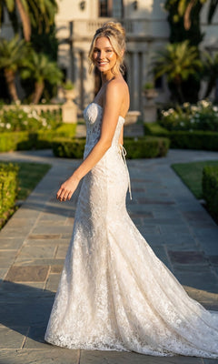 A woman wearing the Juliet JT2667SW long formal dress stands outdoors on a stone path, smiling over her shoulder with greenery and a building in the background.