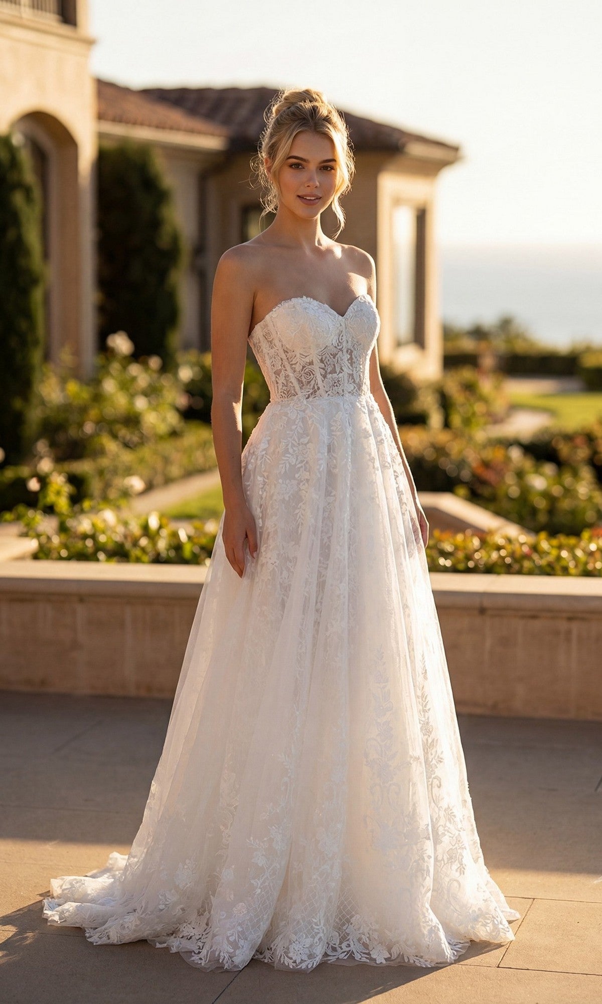 A woman wearing the Juliet JT2670SW Long Prom Dress, featuring a strapless lace design with a sweetheart neckline, stands outdoors in sunlight with a building and garden in the background.
