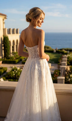 A woman wearing the Juliet JT2670SW long prom dress—featuring a strapless lace design with a sweetheart neckline—stands on a terrace overlooking a garden and the ocean on a sunny day.