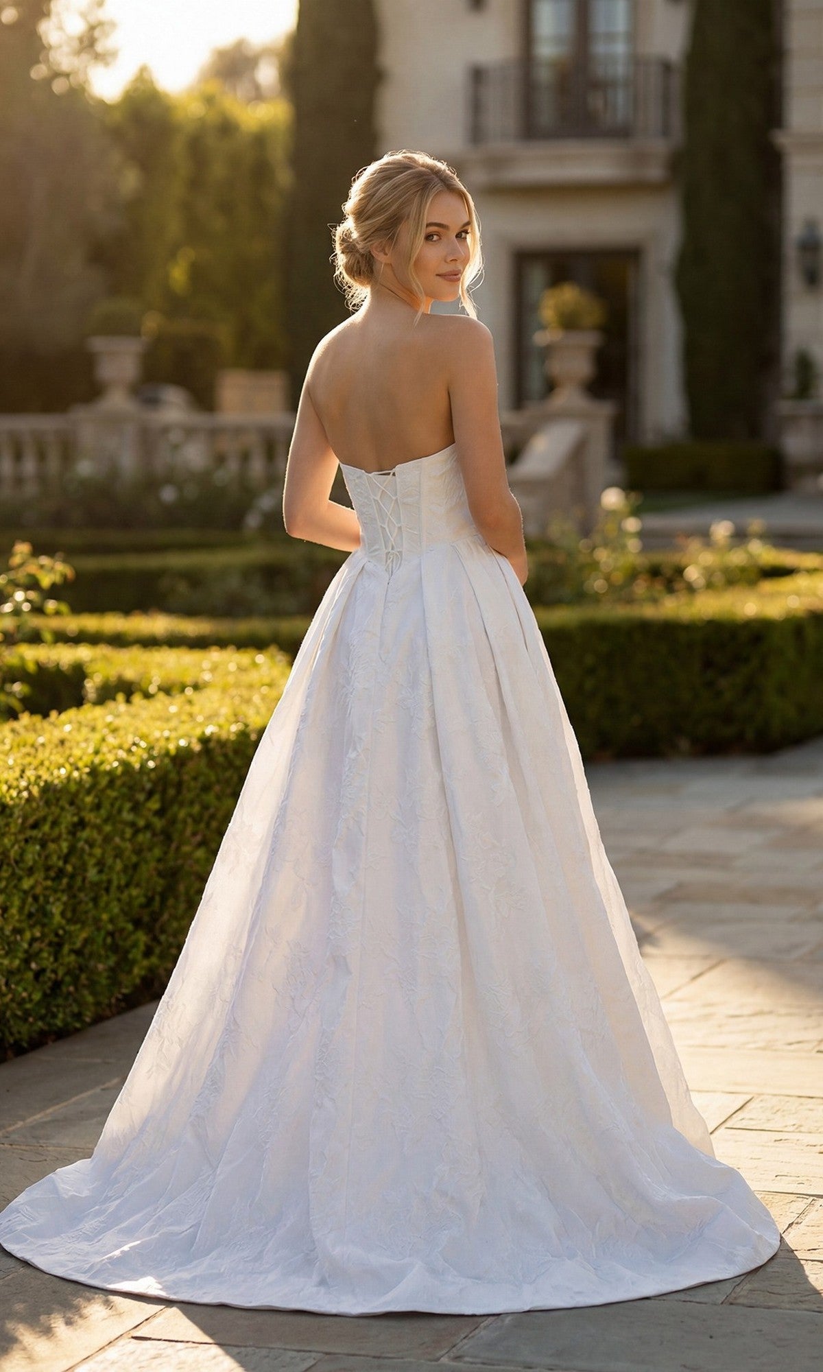 A woman in the Juliet JT2692AW Long Formal Dress stands outdoors on a stone path, looking over her shoulder with a formal building in the background.