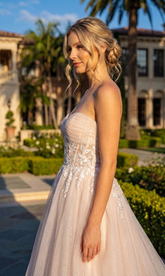 A woman stands outdoors in sunlight wearing the Long Prom Dress: Juliet JT2702AW, a strapless embroidered gown featuring a delicate floral lace corset, with a garden and a large building adorned with arches and palm trees in the background.