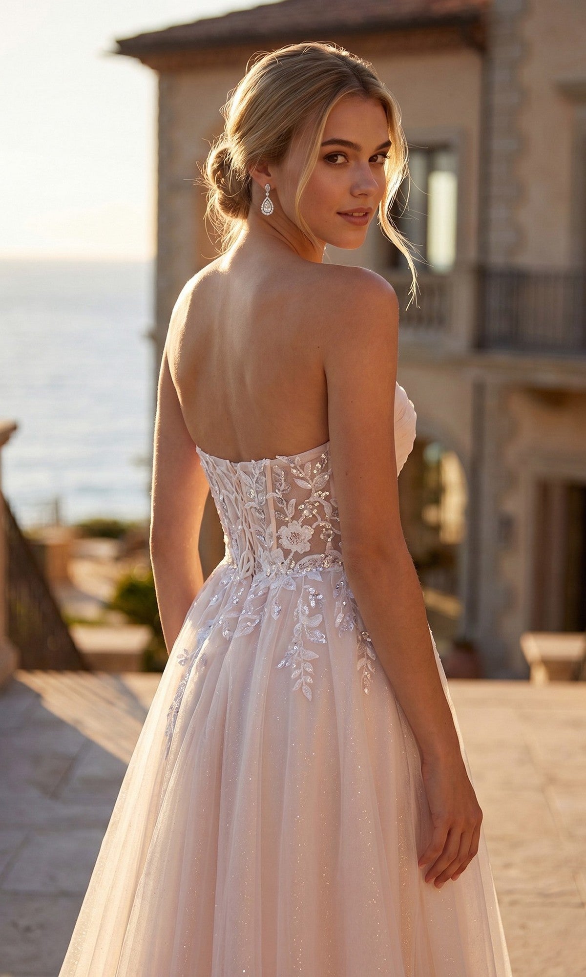 A woman in the Juliet JT2702AW long prom dress stands outdoors, looking over her shoulder. A large house and the sea are visible in the background.