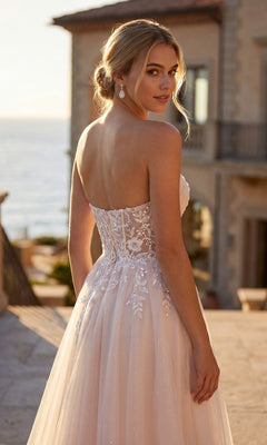 A woman in the Juliet JT2702AW long prom dress stands outdoors, looking over her shoulder. A large house and the sea are visible in the background.