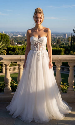 A woman stands outdoors in the off-white Long Prom Dress: Juliet JT2702AW, featuring a floral lace corset bodice and a flowing tulle skirt, smiling with a cityscape and greenery in the background.