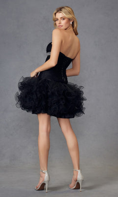 A woman in the Short Black Hoco Dress: Juliet JT705, featuring a strapless neckline and ruffled tulle skirt, with silver high heels, stands facing slightly away against a plain gray background.