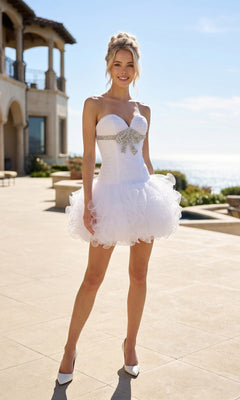 A woman stands outdoors in sunlight wearing the Short Hoco Dress: Juliet JT705 in white, featuring a strapless sweetheart neckline, ruffled tulle skirt, and silver bow bodice, paired with white heels; a large house and ocean are behind her.