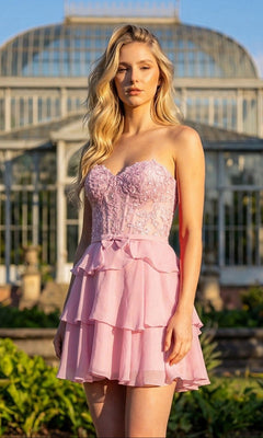 A person with long blonde hair wears the Juliet JT7131G short homecoming dress in Candy Pink, featuring a strapless, layered chiffon A-line skirt, while standing outdoors in front of a large glass conservatory.