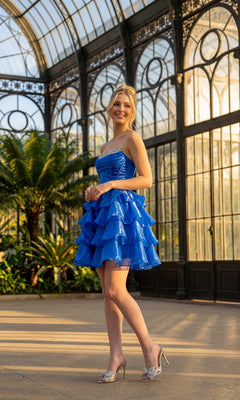 Wearing the Juliet JT7134S short homecoming dress—a vibrant blue, strapless organza design with a ruffle a-line mini skirt—a woman stands smiling in a glass conservatory, surrounded by plants and sunlight streaming through the large windows, her look completed with silver heels.