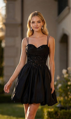 A woman is wearing the Juliet JT7186A short homecoming dress in black, which features a beaded bodice with thin straps and a satin A-line skirt, as she stands outdoors in front of a building and smiles at the camera.