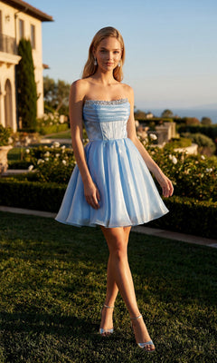 Wearing the Short Homecoming Dress: Juliet JT7187S, a woman stands outdoors on grass near a large building and gardens in the early evening. The strapless design features a delicate corset bodice, and the light blue, knee-length shimmer organza fabric catches the fading sunlight, lending an ethereal glow to her elegant look.