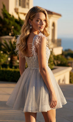 A woman wearing the Juliet JT881 short homecoming dress—a light gray fit-and-flare style with feathered straps—stands outdoors, glancing over her shoulder. Sunlight accentuates her wavy blonde hair as a mansion and lush greenery provide a picturesque backdrop.