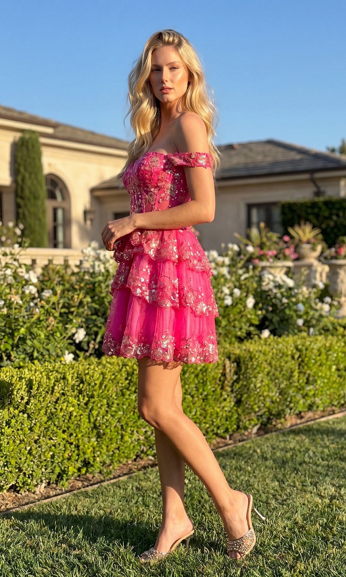 A woman stands on grass in a garden, wearing the Sequin Sheer Corset Homecoming Dress: Juliet JT920K, with flowers and a house in the background.