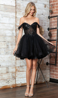 A woman in the Short Black Champagne Corset Prom Dress Juliet JT947L, featuring an off-the-shoulder sequin bodice and a tulle skirt, stands indoors on a wood floor by a brick wall.