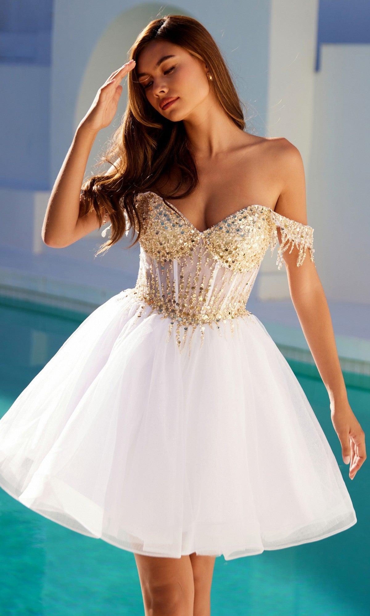 A woman stands by a pool in the Juliet JT947L, a short champagne corset prom dress featuring a white/gold fitted sequin bodice, sheer detailing, and off-the-shoulder tulle.