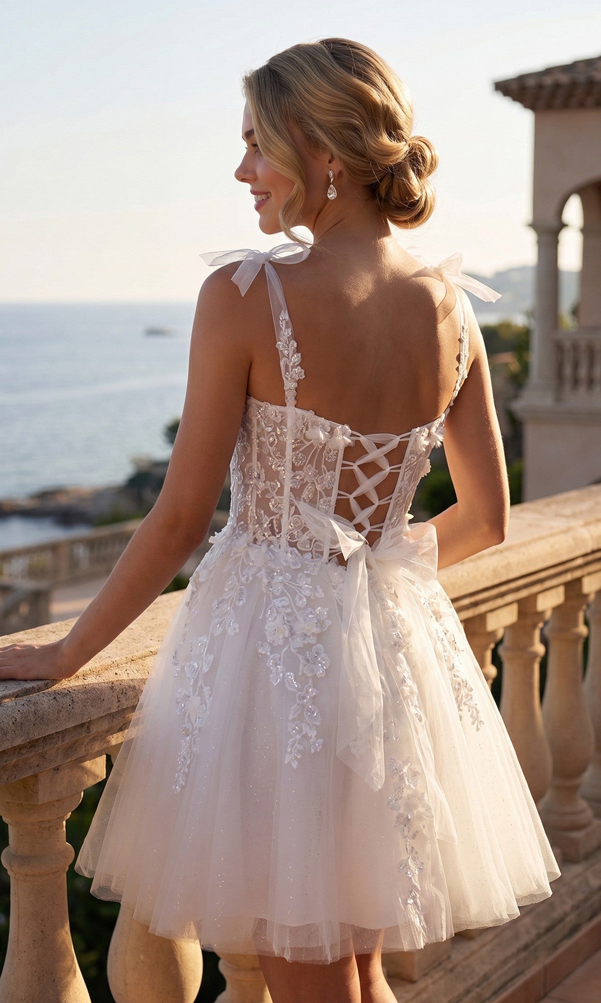 Woman wears the Short White Bow Hoco Dress: Juliet JT972LW, a sleeveless white homecoming dress with floral details and ribbon straps, standing on a balcony overlooking the sea at sunset.