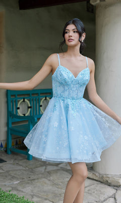 A woman in the Juliet JT985L Short Homecoming Dress, featuring a sleeveless A-line silhouette in Bahama Blue with floral details and a delicate corset bodice, stands outdoors near a turquoise chair and stone wall.