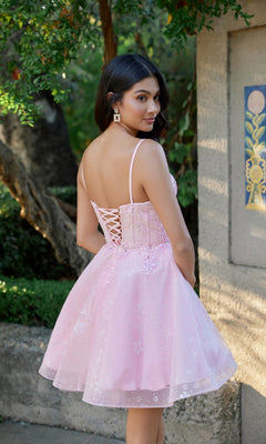 Wearing the Juliet JT985L, a light pink glitter tulle homecoming dress with a lace-up back and corset bodice, a woman stands outdoors near a stone wall and greenery, glancing over her shoulder.