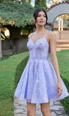 A woman stands outdoors on a stone path beside manicured green hedges, wearing the Juliet JT985L Short Homecoming Dress in lilac—a floral-patterned dress with spaghetti straps and a delicate corset bodice.