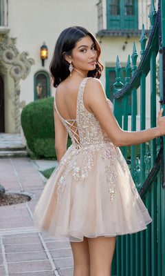 A woman wearing the Juliet JT987N Short Homecoming Dress in champagne, featuring a beautifully embroidered bodice and lace detailing, stands by a green gate outdoors, looking over her shoulder.