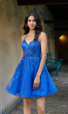 A woman wearing the Juliet JT988N short homecoming dress—a blue, sleeveless, fit-and-flare design with sparkly embellishments—stands on a stone walkway near a wall and a teal bench.