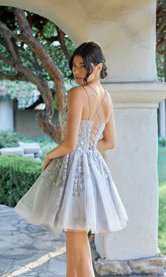 Wearing the Juliet JT988N short homecoming dress—a silver, fit-and-flare style with a lace-up back and floral embellishments—a woman stands outdoors near a stone arch and garden.