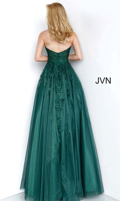 A woman stands facing a white wall wearing the Strapless Long Ball Gown: Jovani 00915—an emerald green, floor-length dress with floral embroidery and embellishments.
