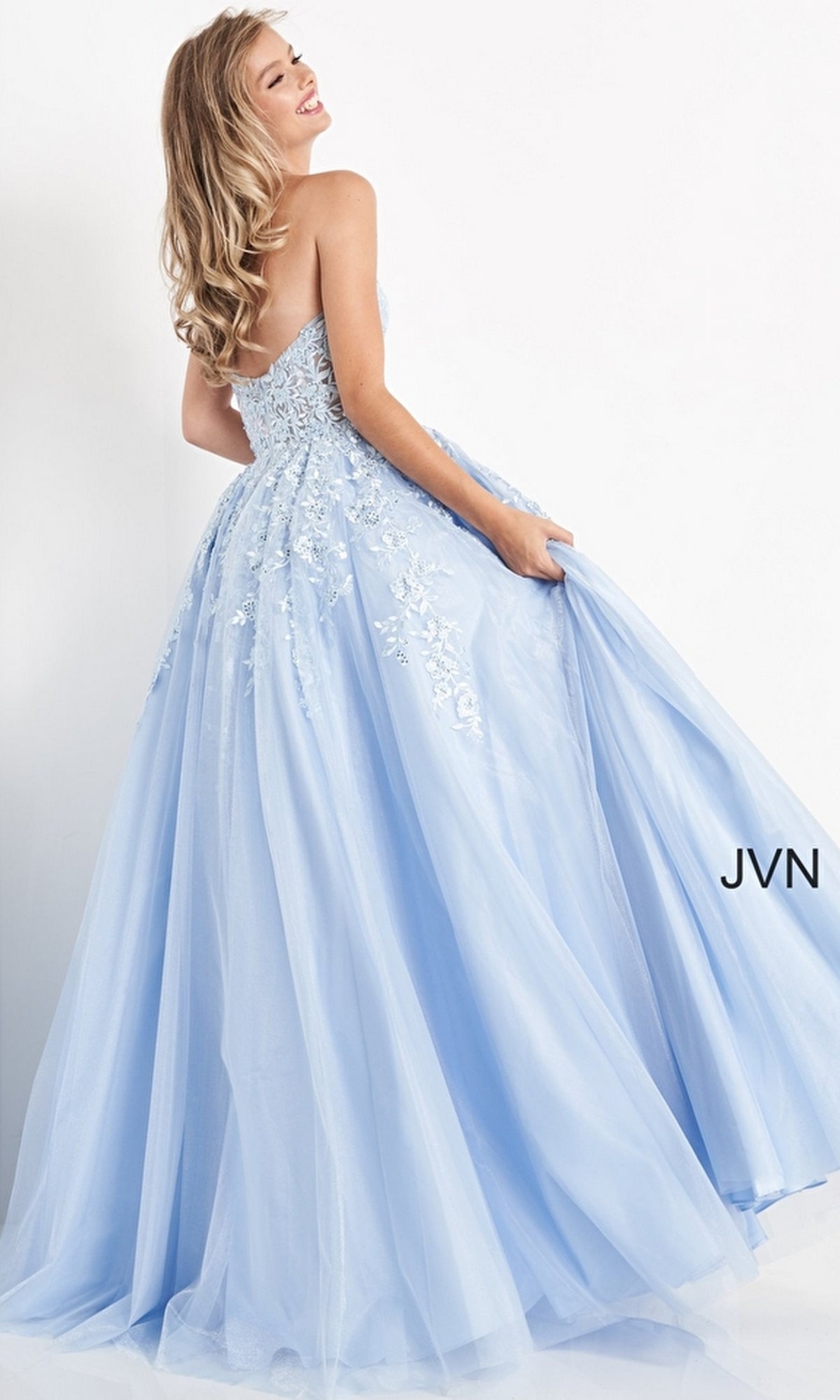A woman smiles while holding her dress, wearing the Strapless Long Ball Gown: Jovani 00915 in light blue with floral lace details. The letters 