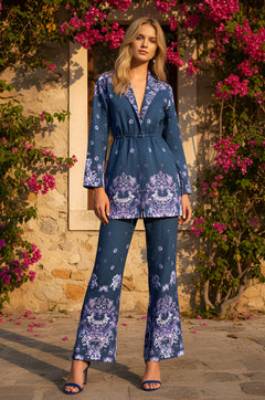 A woman stands outdoors in front of a stone wall and pink flowers, wearing the Summer Paisley-Print Matching Set: June in Regal Paisley, featuring long sleeves, high-rise trousers, and matching heels.