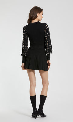 A woman stands with her back to the camera, wearing the Mac Duggal K1007 long sleeve black party dress with grommet sleeve details, paired with black socks and heels, set against a plain light background.